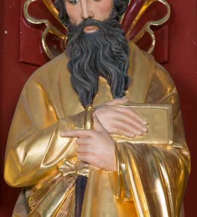 Zdjęcie nr 6: Pełnoplastyczna rzeźba św. Pawła Apostoła została ustawiona na zielonej podstawie na planie kwadratu o ściętych narożach. Święty został ukazany frontalnie w całej postaci. Apostoł ma nieznacznie pochyloną głowę do przodu i ręce skrzyżowane na piersi. Prawą podtrzymuję księgę wspartą o lewe przedramię, trzymając jednocześnie w lewej miecz, skierowany ostrzem ku ziemi. Święty Paweł ma pociągłą twarz okoloną czarnymi włosami i rozdwojoną, długą brodę. Jego spojrzenie skierowane jest w dół. Ma prosty nos, wyraziste oczy i usta. Ubrany jest w przylegającą do ciała tunikę oraz płaszcz narzucony na lewe ramię i podtrzymywany z przodu. Spod tuniki widoczne są bose stopy. W partiach ciała widoczna jest polichromia naturalistyczna. Natomiast szaty i atrybuty są złocone. 