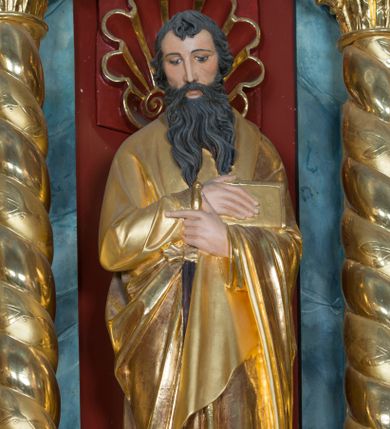 Zdjęcie nr 1: Pełnoplastyczna rzeźba św. Pawła Apostoła została ustawiona na zielonej podstawie na planie kwadratu o ściętych narożach. Święty został ukazany frontalnie w całej postaci. Apostoł ma nieznacznie pochyloną głowę do przodu i ręce skrzyżowane na piersi. Prawą podtrzymuję księgę wspartą o lewe przedramię, trzymając jednocześnie w lewej miecz, skierowany ostrzem ku ziemi. Święty Paweł ma pociągłą twarz okoloną czarnymi włosami i rozdwojoną, długą brodę. Jego spojrzenie skierowane jest w dół. Ma prosty nos, wyraziste oczy i usta. Ubrany jest w przylegającą do ciała tunikę oraz płaszcz narzucony na lewe ramię i podtrzymywany z przodu. Spod tuniki widoczne są bose stopy. W partiach ciała widoczna jest polichromia naturalistyczna. Natomiast szaty i atrybuty są złocone. 