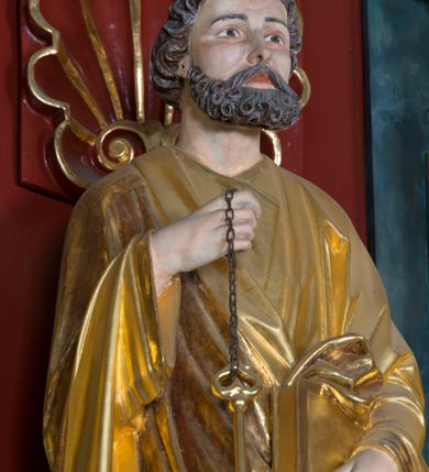 Zdjęcie nr 7: Pełnoplastyczna rzeźba ustawiona na zielonej podstawie na planie kwadratu o ściętych narożach przedstawia św. Piotra Apostoła. Święty został ukazany frontalnie w nieznacznym kontrapoście z prawą ręką uniesioną na wysokość piersi, w której trzyma zawieszone na łańcuszku dwa złote klucze. Lewą ręką podtrzymuje połę płaszcza oraz księgę przyciśnięte do ciała powyżej bioder. Apostoł ma owalną twarz o wyrazistych rysach, otoczoną kędzierzawym, krótkim szpakowatym zarostem i włosami. Jego spojrzenie skierowane jest przed siebie. Ma prosty nos i wydatne usta. Ubrany jest w przylegającą do ciała tunikę oraz płaszcz narzucony na prawe ramię i podtrzymywany z przodu. Spod tuniki widoczne są obute w sandały bose stopy. Obie szaty są silnie drapowane. W partiach ciała widoczna jest naturalistyczna polichromia. Natomiast ubranie i atrybuty zostały wyzłocone.