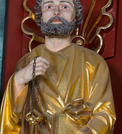 Zdjęcie nr 6: Pełnoplastyczna rzeźba ustawiona na zielonej podstawie na planie kwadratu o ściętych narożach przedstawia św. Piotra Apostoła. Święty został ukazany frontalnie w nieznacznym kontrapoście z prawą ręką uniesioną na wysokość piersi, w której trzyma zawieszone na łańcuszku dwa złote klucze. Lewą ręką podtrzymuje połę płaszcza oraz księgę przyciśnięte do ciała powyżej bioder. Apostoł ma owalną twarz o wyrazistych rysach, otoczoną kędzierzawym, krótkim szpakowatym zarostem i włosami. Jego spojrzenie skierowane jest przed siebie. Ma prosty nos i wydatne usta. Ubrany jest w przylegającą do ciała tunikę oraz płaszcz narzucony na prawe ramię i podtrzymywany z przodu. Spod tuniki widoczne są obute w sandały bose stopy. Obie szaty są silnie drapowane. W partiach ciała widoczna jest naturalistyczna polichromia. Natomiast ubranie i atrybuty zostały wyzłocone.