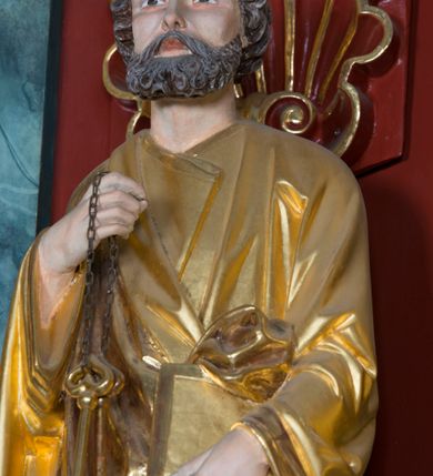 Zdjęcie nr 5: Pełnoplastyczna rzeźba ustawiona na zielonej podstawie na planie kwadratu o ściętych narożach przedstawia św. Piotra Apostoła. Święty został ukazany frontalnie w nieznacznym kontrapoście z prawą ręką uniesioną na wysokość piersi, w której trzyma zawieszone na łańcuszku dwa złote klucze. Lewą ręką podtrzymuje połę płaszcza oraz księgę przyciśnięte do ciała powyżej bioder. Apostoł ma owalną twarz o wyrazistych rysach, otoczoną kędzierzawym, krótkim szpakowatym zarostem i włosami. Jego spojrzenie skierowane jest przed siebie. Ma prosty nos i wydatne usta. Ubrany jest w przylegającą do ciała tunikę oraz płaszcz narzucony na prawe ramię i podtrzymywany z przodu. Spod tuniki widoczne są obute w sandały bose stopy. Obie szaty są silnie drapowane. W partiach ciała widoczna jest naturalistyczna polichromia. Natomiast ubranie i atrybuty zostały wyzłocone.