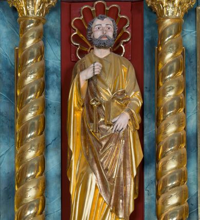 Zdjęcie nr 2: Pełnoplastyczna rzeźba ustawiona na zielonej podstawie na planie kwadratu o ściętych narożach przedstawia św. Piotra Apostoła. Święty został ukazany frontalnie w nieznacznym kontrapoście z prawą ręką uniesioną na wysokość piersi, w której trzyma zawieszone na łańcuszku dwa złote klucze. Lewą ręką podtrzymuje połę płaszcza oraz księgę przyciśnięte do ciała powyżej bioder. Apostoł ma owalną twarz o wyrazistych rysach, otoczoną kędzierzawym, krótkim szpakowatym zarostem i włosami. Jego spojrzenie skierowane jest przed siebie. Ma prosty nos i wydatne usta. Ubrany jest w przylegającą do ciała tunikę oraz płaszcz narzucony na prawe ramię i podtrzymywany z przodu. Spod tuniki widoczne są obute w sandały bose stopy. Obie szaty są silnie drapowane. W partiach ciała widoczna jest naturalistyczna polichromia. Natomiast ubranie i atrybuty zostały wyzłocone.