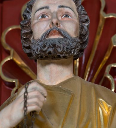 Zdjęcie nr 8: Pełnoplastyczna rzeźba ustawiona na zielonej podstawie na planie kwadratu o ściętych narożach przedstawia św. Piotra Apostoła. Święty został ukazany frontalnie w nieznacznym kontrapoście z prawą ręką uniesioną na wysokość piersi, w której trzyma zawieszone na łańcuszku dwa złote klucze. Lewą ręką podtrzymuje połę płaszcza oraz księgę przyciśnięte do ciała powyżej bioder. Apostoł ma owalną twarz o wyrazistych rysach, otoczoną kędzierzawym, krótkim szpakowatym zarostem i włosami. Jego spojrzenie skierowane jest przed siebie. Ma prosty nos i wydatne usta. Ubrany jest w przylegającą do ciała tunikę oraz płaszcz narzucony na prawe ramię i podtrzymywany z przodu. Spod tuniki widoczne są obute w sandały bose stopy. Obie szaty są silnie drapowane. W partiach ciała widoczna jest naturalistyczna polichromia. Natomiast ubranie i atrybuty zostały wyzłocone.