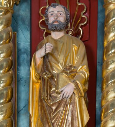 Zdjęcie nr 1: Pełnoplastyczna rzeźba ustawiona na zielonej podstawie na planie kwadratu o ściętych narożach przedstawia św. Piotra Apostoła. Święty został ukazany frontalnie w nieznacznym kontrapoście z prawą ręką uniesioną na wysokość piersi, w której trzyma zawieszone na łańcuszku dwa złote klucze. Lewą ręką podtrzymuje połę płaszcza oraz księgę przyciśnięte do ciała powyżej bioder. Apostoł ma owalną twarz o wyrazistych rysach, otoczoną kędzierzawym, krótkim szpakowatym zarostem i włosami. Jego spojrzenie skierowane jest przed siebie. Ma prosty nos i wydatne usta. Ubrany jest w przylegającą do ciała tunikę oraz płaszcz narzucony na prawe ramię i podtrzymywany z przodu. Spod tuniki widoczne są obute w sandały bose stopy. Obie szaty są silnie drapowane. W partiach ciała widoczna jest naturalistyczna polichromia. Natomiast ubranie i atrybuty zostały wyzłocone.