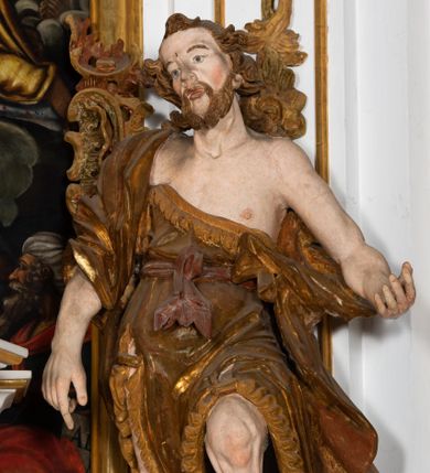 Zdjęcie nr 1: Ustawiona na cokole o nieregularnym planie, pełnoplastyczna rzeźba drążona, ukazuje św. Jana Chrzciciela. Figura zwrócona jest do widza frontalnie, w kontrapoście, z głową lekko przechyloną w prawo, o silnym esowatym wygięciu ciała. Święty prawą rękę opuszcza wzdłuż ciała. Jego dłoń zdaje się wskazywać coś na dole. Natomiast lewą rękę ma wyciągniętą w bok. Być może pierwotnie podtrzymywał nią księgę z przedstawieniem Baranka. Jan Chrzciciel został ukazany jako dojrzały mężczyzna. Jego ciało jest szczupłe o wyraźnie podkreślonej muskulaturze rąk i nóg oraz mostku i obojczykach klatki piersiowej. Ma szczupłą, pociągłą twarz, o silnie podkreślonych kościach policzkowych, łukach brwiowych i silnie zadartym, wąskim nosie z wysuniętą do przodu górną wargą, oczy niewielkie, okrągłe, usta pełne i lekko rozchylone. Twarz okala krótki zarost skręcony w dwa pukle oraz włosy układające się w rozwiane, silne skręcone pukle na czubku głowy i po jej bokach. Święty ubrany jest w silnie przylegającą do ciała, lekko marszczoną melotę przewiązaną w pasie szarfą, odsłaniającą połowę torsu oraz nogi. Na prawe ramię ma narzucony silnie drapowany płaszcz, opadający z tyłu i na lewy boku postaci. W odsłoniętych partiach ciała polichromia rzeźby jest naturalistyczna, szaty złocone natomiast szarfa w pasie srebrzona.