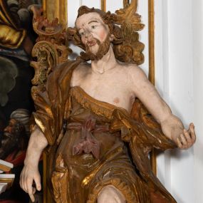 Zdjęcie nr 1: Ustawiona na cokole o nieregularnym planie, pełnoplastyczna rzeźba drążona, ukazuje św. Jana Chrzciciela. Figura zwrócona jest do widza frontalnie, w kontrapoście, z głową lekko przechyloną w prawo, o silnym esowatym wygięciu ciała. Święty prawą rękę opuszcza wzdłuż ciała. Jego dłoń zdaje się wskazywać coś na dole. Natomiast lewą rękę ma wyciągniętą w bok. Być może pierwotnie podtrzymywał nią księgę z przedstawieniem Baranka. Jan Chrzciciel został ukazany jako dojrzały mężczyzna. Jego ciało jest szczupłe o wyraźnie podkreślonej muskulaturze rąk i nóg oraz mostku i obojczykach klatki piersiowej. Ma szczupłą, pociągłą twarz, o silnie podkreślonych kościach policzkowych, łukach brwiowych i silnie zadartym, wąskim nosie z wysuniętą do przodu górną wargą, oczy niewielkie, okrągłe, usta pełne i lekko rozchylone. Twarz okala krótki zarost skręcony w dwa pukle oraz włosy układające się w rozwiane, silne skręcone pukle na czubku głowy i po jej bokach. Święty ubrany jest w silnie przylegającą do ciała, lekko marszczoną melotę przewiązaną w pasie szarfą, odsłaniającą połowę torsu oraz nogi. Na prawe ramię ma narzucony silnie drapowany płaszcz, opadający z tyłu i na lewy boku postaci. W odsłoniętych partiach ciała polichromia rzeźby jest naturalistyczna, szaty złocone natomiast szarfa w pasie srebrzona.