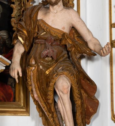 Zdjęcie nr 2: Ustawiona na cokole o nieregularnym planie, pełnoplastyczna rzeźba drążona, ukazuje św. Jana Chrzciciela. Figura zwrócona jest do widza frontalnie, w kontrapoście, z głową lekko przechyloną w prawo, o silnym esowatym wygięciu ciała. Święty prawą rękę opuszcza wzdłuż ciała. Jego dłoń zdaje się wskazywać coś na dole. Natomiast lewą rękę ma wyciągniętą w bok. Być może pierwotnie podtrzymywał nią księgę z przedstawieniem Baranka. Jan Chrzciciel został ukazany jako dojrzały mężczyzna. Jego ciało jest szczupłe o wyraźnie podkreślonej muskulaturze rąk i nóg oraz mostku i obojczykach klatki piersiowej. Ma szczupłą, pociągłą twarz, o silnie podkreślonych kościach policzkowych, łukach brwiowych i silnie zadartym, wąskim nosie z wysuniętą do przodu górną wargą, oczy niewielkie, okrągłe, usta pełne i lekko rozchylone. Twarz okala krótki zarost skręcony w dwa pukle oraz włosy układające się w rozwiane, silne skręcone pukle na czubku głowy i po jej bokach. Święty ubrany jest w silnie przylegającą do ciała, lekko marszczoną melotę przewiązaną w pasie szarfą, odsłaniającą połowę torsu oraz nogi. Na prawe ramię ma narzucony silnie drapowany płaszcz, opadający z tyłu i na lewy boku postaci. W odsłoniętych partiach ciała polichromia rzeźby jest naturalistyczna, szaty złocone natomiast szarfa w pasie srebrzona.
