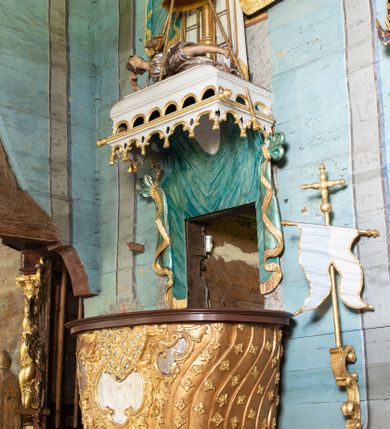 Zdjęcie nr 1: Ambona uzyskała kształt łodzi zawieszonej na północnej ścianie prezbiterium przy łuku tęczowym. Wejście na ambonę prowadzi z zakrystii. Jej kosz uzyskał owalną formę rozszerzającą się ku górze. W dolnej partii do jednej trzeciej wysokości widoczne są rzeźbione przedstawienia wody ze stworami morskimi (delfiny i ślimaki). Natomiast powyżej kosz dekorowany jest od frontu trzema rokokowymi kartuszami oraz fragmentem wzorzystej tkaniny, flankowanymi kilkoma parami falistych pasów zdobionych rozetami. Zaplecek ambony uzyskał kształt rzeźbionej, opadającej tkaniny podwiązanej po bokach w kolorze opalizującej zieleni, lamowanej złotymi frędzlami. Ponad amboną zawieszono baldachim na planie prostokąta, z profilowanymi bokami, z ciągiem ażurowych arkadek u dołu. Przy dolnej krawędzi zdobi go lambrekin ze złotymi chwostami. Na baldachimie leży postać wspierająca głowę na prawej ręce. Lewą rękę ma uniesioną do góry. Postać o krótkich włosach otaczających twarz o jasnej karnacji z drobnymi rysami, ubrana jest w srebrzystą tunikę przewiązaną w pasie i odsłaniającą lewą nogę na wysokości kolana. Ponad baldachimem wznosi się maszt z żaglem, którego liny opasane są dwiema złotymi koronami o floralnych sterczynach. Maszt flankują dwie rozwiane tkaniny w kolorze opalizującej zieleni, lamowane złotymi frędzlami, wiszące na złotych drążkach o puklowanych zakończeniach. Wieńczy go promienista gloria z gołębicą Ducha Świętego w otoczeniu obłoków. Obok kosza ambony do ściany przytwierdzono złocony, wolutowy wspornik z proporcem zwieńczonym krzyżem.