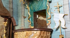 Zdjęcie nr 1: Ambona uzyskała kształt łodzi zawieszonej na północnej ścianie prezbiterium przy łuku tęczowym. Wejście na ambonę prowadzi z zakrystii. Jej kosz uzyskał owalną formę rozszerzającą się ku górze. W dolnej partii do jednej trzeciej wysokości widoczne są rzeźbione przedstawienia wody ze stworami morskimi (delfiny i ślimaki). Natomiast powyżej kosz dekorowany jest od frontu trzema rokokowymi kartuszami oraz fragmentem wzorzystej tkaniny, flankowanymi kilkoma parami falistych pasów zdobionych rozetami. Zaplecek ambony uzyskał kształt rzeźbionej, opadającej tkaniny podwiązanej po bokach w kolorze opalizującej zieleni, lamowanej złotymi frędzlami. Ponad amboną zawieszono baldachim na planie prostokąta, z profilowanymi bokami, z ciągiem ażurowych arkadek u dołu. Przy dolnej krawędzi zdobi go lambrekin ze złotymi chwostami. Na baldachimie leży postać wspierająca głowę na prawej ręce. Lewą rękę ma uniesioną do góry. Postać o krótkich włosach otaczających twarz o jasnej karnacji z drobnymi rysami, ubrana jest w srebrzystą tunikę przewiązaną w pasie i odsłaniającą lewą nogę na wysokości kolana. Ponad baldachimem wznosi się maszt z żaglem, którego liny opasane są dwiema złotymi koronami o floralnych sterczynach. Maszt flankują dwie rozwiane tkaniny w kolorze opalizującej zieleni, lamowane złotymi frędzlami, wiszące na złotych drążkach o puklowanych zakończeniach. Wieńczy go promienista gloria z gołębicą Ducha Świętego w otoczeniu obłoków. Obok kosza ambony do ściany przytwierdzono złocony, wolutowy wspornik z proporcem zwieńczonym krzyżem.