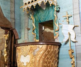 Zdjęcie nr 1: Ambona uzyskała kształt łodzi zawieszonej na północnej ścianie prezbiterium przy łuku tęczowym. Wejście na ambonę prowadzi z zakrystii. Jej kosz uzyskał owalną formę rozszerzającą się ku górze. W dolnej partii do jednej trzeciej wysokości widoczne są rzeźbione przedstawienia wody ze stworami morskimi (delfiny i ślimaki). Natomiast powyżej kosz dekorowany jest od frontu trzema rokokowymi kartuszami oraz fragmentem wzorzystej tkaniny, flankowanymi kilkoma parami falistych pasów zdobionych rozetami. Zaplecek ambony uzyskał kształt rzeźbionej, opadającej tkaniny podwiązanej po bokach w kolorze opalizującej zieleni, lamowanej złotymi frędzlami. Ponad amboną zawieszono baldachim na planie prostokąta, z profilowanymi bokami, z ciągiem ażurowych arkadek u dołu. Przy dolnej krawędzi zdobi go lambrekin ze złotymi chwostami. Na baldachimie leży postać wspierająca głowę na prawej ręce. Lewą rękę ma uniesioną do góry. Postać o krótkich włosach otaczających twarz o jasnej karnacji z drobnymi rysami, ubrana jest w srebrzystą tunikę przewiązaną w pasie i odsłaniającą lewą nogę na wysokości kolana. Ponad baldachimem wznosi się maszt z żaglem, którego liny opasane są dwiema złotymi koronami o floralnych sterczynach. Maszt flankują dwie rozwiane tkaniny w kolorze opalizującej zieleni, lamowane złotymi frędzlami, wiszące na złotych drążkach o puklowanych zakończeniach. Wieńczy go promienista gloria z gołębicą Ducha Świętego w otoczeniu obłoków. Obok kosza ambony do ściany przytwierdzono złocony, wolutowy wspornik z proporcem zwieńczonym krzyżem.