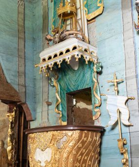 Zdjęcie nr 1: Ambona uzyskała kształt łodzi zawieszonej na północnej ścianie prezbiterium przy łuku tęczowym. Wejście na ambonę prowadzi z zakrystii. Jej kosz uzyskał owalną formę rozszerzającą się ku górze. W dolnej partii do jednej trzeciej wysokości widoczne są rzeźbione przedstawienia wody ze stworami morskimi (delfiny i ślimaki). Natomiast powyżej kosz dekorowany jest od frontu trzema rokokowymi kartuszami oraz fragmentem wzorzystej tkaniny, flankowanymi kilkoma parami falistych pasów zdobionych rozetami. Zaplecek ambony uzyskał kształt rzeźbionej, opadającej tkaniny podwiązanej po bokach w kolorze opalizującej zieleni, lamowanej złotymi frędzlami. Ponad amboną zawieszono baldachim na planie prostokąta, z profilowanymi bokami, z ciągiem ażurowych arkadek u dołu. Przy dolnej krawędzi zdobi go lambrekin ze złotymi chwostami. Na baldachimie leży postać wspierająca głowę na prawej ręce. Lewą rękę ma uniesioną do góry. Postać o krótkich włosach otaczających twarz o jasnej karnacji z drobnymi rysami, ubrana jest w srebrzystą tunikę przewiązaną w pasie i odsłaniającą lewą nogę na wysokości kolana. Ponad baldachimem wznosi się maszt z żaglem, którego liny opasane są dwiema złotymi koronami o floralnych sterczynach. Maszt flankują dwie rozwiane tkaniny w kolorze opalizującej zieleni, lamowane złotymi frędzlami, wiszące na złotych drążkach o puklowanych zakończeniach. Wieńczy go promienista gloria z gołębicą Ducha Świętego w otoczeniu obłoków. Obok kosza ambony do ściany przytwierdzono złocony, wolutowy wspornik z proporcem zwieńczonym krzyżem.