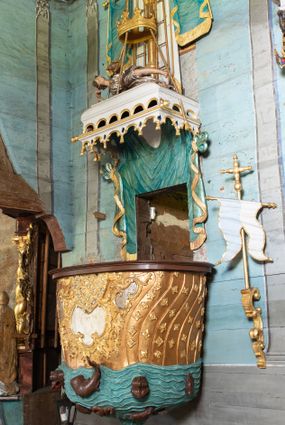 Zdjęcie nr 1: Ambona uzyskała kształt łodzi zawieszonej na północnej ścianie prezbiterium przy łuku tęczowym. Wejście na ambonę prowadzi z zakrystii. Jej kosz uzyskał owalną formę rozszerzającą się ku górze. W dolnej partii do jednej trzeciej wysokości widoczne są rzeźbione przedstawienia wody ze stworami morskimi (delfiny i ślimaki). Natomiast powyżej kosz dekorowany jest od frontu trzema rokokowymi kartuszami oraz fragmentem wzorzystej tkaniny, flankowanymi kilkoma parami falistych pasów zdobionych rozetami. Zaplecek ambony uzyskał kształt rzeźbionej, opadającej tkaniny podwiązanej po bokach w kolorze opalizującej zieleni, lamowanej złotymi frędzlami. Ponad amboną zawieszono baldachim na planie prostokąta, z profilowanymi bokami, z ciągiem ażurowych arkadek u dołu. Przy dolnej krawędzi zdobi go lambrekin ze złotymi chwostami. Na baldachimie leży postać wspierająca głowę na prawej ręce. Lewą rękę ma uniesioną do góry. Postać o krótkich włosach otaczających twarz o jasnej karnacji z drobnymi rysami, ubrana jest w srebrzystą tunikę przewiązaną w pasie i odsłaniającą lewą nogę na wysokości kolana. Ponad baldachimem wznosi się maszt z żaglem, którego liny opasane są dwiema złotymi koronami o floralnych sterczynach. Maszt flankują dwie rozwiane tkaniny w kolorze opalizującej zieleni, lamowane złotymi frędzlami, wiszące na złotych drążkach o puklowanych zakończeniach. Wieńczy go promienista gloria z gołębicą Ducha Świętego w otoczeniu obłoków. Obok kosza ambony do ściany przytwierdzono złocony, wolutowy wspornik z proporcem zwieńczonym krzyżem.