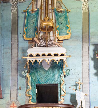 Zdjęcie nr 2: Ambona uzyskała kształt łodzi zawieszonej na północnej ścianie prezbiterium przy łuku tęczowym. Wejście na ambonę prowadzi z zakrystii. Jej kosz uzyskał owalną formę rozszerzającą się ku górze. W dolnej partii do jednej trzeciej wysokości widoczne są rzeźbione przedstawienia wody ze stworami morskimi (delfiny i ślimaki). Natomiast powyżej kosz dekorowany jest od frontu trzema rokokowymi kartuszami oraz fragmentem wzorzystej tkaniny, flankowanymi kilkoma parami falistych pasów zdobionych rozetami. Zaplecek ambony uzyskał kształt rzeźbionej, opadającej tkaniny podwiązanej po bokach w kolorze opalizującej zieleni, lamowanej złotymi frędzlami. Ponad amboną zawieszono baldachim na planie prostokąta, z profilowanymi bokami, z ciągiem ażurowych arkadek u dołu. Przy dolnej krawędzi zdobi go lambrekin ze złotymi chwostami. Na baldachimie leży postać wspierająca głowę na prawej ręce. Lewą rękę ma uniesioną do góry. Postać o krótkich włosach otaczających twarz o jasnej karnacji z drobnymi rysami, ubrana jest w srebrzystą tunikę przewiązaną w pasie i odsłaniającą lewą nogę na wysokości kolana. Ponad baldachimem wznosi się maszt z żaglem, którego liny opasane są dwiema złotymi koronami o floralnych sterczynach. Maszt flankują dwie rozwiane tkaniny w kolorze opalizującej zieleni, lamowane złotymi frędzlami, wiszące na złotych drążkach o puklowanych zakończeniach. Wieńczy go promienista gloria z gołębicą Ducha Świętego w otoczeniu obłoków. Obok kosza ambony do ściany przytwierdzono złocony, wolutowy wspornik z proporcem zwieńczonym krzyżem.