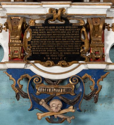 Zdjęcie nr 9: Epitafium uzyskało formę jednokondygnacyjnej, trójosiowej struktury architektonicznej z podstawą i zwieńczeniem, flankowanej po bokach fantazyjnymi uszakami. Część środkową tworzą trzy aedicule rozdzielone dwiema złoconymi kolumnami o korynckich kapitelach i trzonach dekorowanych festonami, a w dolnych partiach pierścieniami. Kolumny podtrzymują pełne belkowanie przechodzące przez całą szerokości kondygnacji, wyłamane nad osiami bocznymi. Same zaś stoją na cokole, który stanowi równocześnie wyłamany gzyms dla podstawy. Pole osi środkowej otrzymało kształt aediculi w formie stojącego prostokąta ze schodkowym zamknięciem u góry, wypełnionym ażurowym, złoconym maswerkiem. Jej wnętrze wypełnia płaskorzeźbione słońce pośród chmur na malowanym tle nieba. Poniżej ukazano panoramę Jerozolimy. Przed aediculą ustawiono na postumencie drewniany krzyż. Na osiach bocznych znalazły się nisze zwieńczone złotymi muszlami oraz złoconą, rytą ornamentyką z motywem czteroliścia. Pierwotnie znajdowały się tu rzeźby Mojżesza i Jana Chrzciciela. Całość po bokach flankują fantazyjne uszaki utworzone ornamentu rollwerkowego oraz z widocznych z profilu półpostaci maurów podtrzymujących belkowanie. Pole uszaków wypełniają pionowo ułożone owalne kartusze o czerwonych polach, stanowiące tło dla dwóch par tarczy herbowych z klejnotami. Z lewej umieszczono herb Dołęga, a z prawej herb Jastrzębiec (bez klejnotu). Po zewnętrznych stronach na belkowaniu ustawiono dwa półsiedzące uskrzydlone anioły (zachował się tylko anioł z prawej strony). Jednocześnie postacie aniołów flankują zwieńczenie epitafium, które tworzy aedicula wypełniona złotą muszlą oraz rytym złoconym ornamentem z motywem czteroliścia. Flankują ją dwa pilastry podtrzymujące wyłamane belkowanie na ich osi. W aediculi ustawiono pełnoplastyczną rzeźbę stojącej kobiety. Została ona ukazana w wydatnym kontrapoście z tułowiem skręconym w lewo, z głową ujętą w trzech czwartych w prawo. Kobieta w podniesionej prawej ręce trzyma srebrny kielich, natomiast w opuszczonej i wspartej o wysuniętą tanecznie lewą nogę otwartą księgę. Postać ma owalną twarz o jasnej karnacji i rumianych policzkach z prostym nosem i wydatnymi ustami. Jej głowę otaczają falujące, brązowe włosy opadające na plecy i  odsłaniające uszy. Ubrana jest w złoconą, mocno drapowaną suknię, która odsłania wysuniętą nogę. Po bokach pilastrów widoczne są dwa uszaki. Na szczycie zwieńczenia dwa pulchne putta prezentują chustę Weroniki, która została równocześnie przytwierdzona do wysokiego impostu wspierającego ażurowy, złocony kwiaton. Na chuście wyobrażono płaskorzeźbioną głowę Chrystusa w cierniowej koronie. Zbawiciel ma owalną twarz z szeroko otwartymi oczami, wydatnym nosem i półotwartymi ustami. Otaczają ją kręcone brązowe włosy oraz podobna, krótka broda. Podstawę epitafium tworzy wykrojona po bokach struktura, w której centrum umieszczono fantazyjnie wyciętą drewnianą, czarną tablicę ze złotym łacińskim napisem "O HOMO FVI QVOD ES SVM QVOD ERIS / NVNC DE(M)VM VIVERE CO(E)PI, MORI DESII, VITA MIHI / PRIOR MORTE(M), MORS PRIA(M) VITA(M) SECV(N)DA(M), QVAE NU(N) QVA(M) / EST I(N)TERI TVRA DEDIT, MO(R)TALITATES  MEOR, HO(C) MIHI / EST ET VXORI MO(S) VENTV(S) POSVI, VIPOST QV(E)ALETHIVERA DVCEDI / NELVSOS, ET CARNIS EXVVIIS SO(L)VTOS  ABHOC PELAG  SECVRA / GLAVSTRA SVB IRE IVABIT, HIC MORTALE DIMIDIVM N(OST)RI, IMMORTA/LE(M) EXPECT ET IM(M)ORTALITATEM, E QVOS VITA CONIVCTOS TENVIT / MORS IDIVISOS CVSTODIRET, SI PIVS ES, PIAS PRECES ADDE, ET / HIC HV(M)O SVB HVMO QVIETA[M], ANIMAE IN COELESTI PATRIA / LETAM  QVIETEM APPRECARE "NICOLAVS CORNITIVS DE / MAGNA POREMBA ET BEATA DE MIROW CONSOR/TES SE(M)PER VAIESL[?]" / A(NNO) D(OMINI) 1611 P(ATRONVS) P(OSVIT)". Tablicę umieszczono w kartuszu rollwerkowym pomiędzy dwiema konsolami zdobionymi pierwotnie uskrzydlonymi główkami aniołków, podtrzymującymi belkowanie z krzyżem. Poniżej tablicy zastosowano odwrócony łuk odcinkowy z podwieszonym kartuszem rollwerkowym zawieszonym na festonach. W jego wnętrzu przedstawiono tabliczkę z napisem „VITA FUGIT VT HORA”, a po niżej trupią czaszkę ze skrzyżowanymi piszczelami.  
