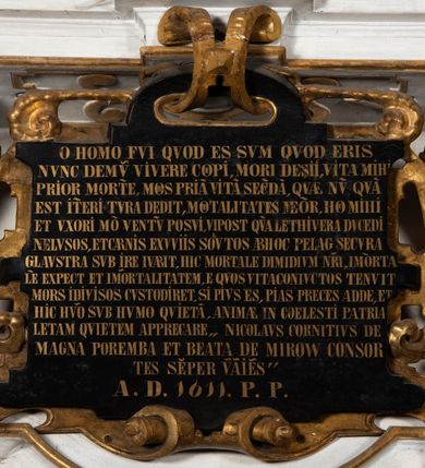 Zdjęcie nr 5: Epitafium uzyskało formę jednokondygnacyjnej, trójosiowej struktury architektonicznej z podstawą i zwieńczeniem, flankowanej po bokach fantazyjnymi uszakami. Część środkową tworzą trzy aedicule rozdzielone dwiema złoconymi kolumnami o korynckich kapitelach i trzonach dekorowanych festonami, a w dolnych partiach pierścieniami. Kolumny podtrzymują pełne belkowanie przechodzące przez całą szerokości kondygnacji, wyłamane nad osiami bocznymi. Same zaś stoją na cokole, który stanowi równocześnie wyłamany gzyms dla podstawy. Pole osi środkowej otrzymało kształt aediculi w formie stojącego prostokąta ze schodkowym zamknięciem u góry, wypełnionym ażurowym, złoconym maswerkiem. Jej wnętrze wypełnia płaskorzeźbione słońce pośród chmur na malowanym tle nieba. Poniżej ukazano panoramę Jerozolimy. Przed aediculą ustawiono na postumencie drewniany krzyż. Na osiach bocznych znalazły się nisze zwieńczone złotymi muszlami oraz złoconą, rytą ornamentyką z motywem czteroliścia. Pierwotnie znajdowały się tu rzeźby Mojżesza i Jana Chrzciciela. Całość po bokach flankują fantazyjne uszaki utworzone ornamentu rollwerkowego oraz z widocznych z profilu półpostaci maurów podtrzymujących belkowanie. Pole uszaków wypełniają pionowo ułożone owalne kartusze o czerwonych polach, stanowiące tło dla dwóch par tarczy herbowych z klejnotami. Z lewej umieszczono herb Dołęga, a z prawej herb Jastrzębiec (bez klejnotu). Po zewnętrznych stronach na belkowaniu ustawiono dwa półsiedzące uskrzydlone anioły (zachował się tylko anioł z prawej strony). Jednocześnie postacie aniołów flankują zwieńczenie epitafium, które tworzy aedicula wypełniona złotą muszlą oraz rytym złoconym ornamentem z motywem czteroliścia. Flankują ją dwa pilastry podtrzymujące wyłamane belkowanie na ich osi. W aediculi ustawiono pełnoplastyczną rzeźbę stojącej kobiety. Została ona ukazana w wydatnym kontrapoście z tułowiem skręconym w lewo, z głową ujętą w trzech czwartych w prawo. Kobieta w podniesionej prawej ręce trzyma srebrny kielich, natomiast w opuszczonej i wspartej o wysuniętą tanecznie lewą nogę otwartą księgę. Postać ma owalną twarz o jasnej karnacji i rumianych policzkach z prostym nosem i wydatnymi ustami. Jej głowę otaczają falujące, brązowe włosy opadające na plecy i  odsłaniające uszy. Ubrana jest w złoconą, mocno drapowaną suknię, która odsłania wysuniętą nogę. Po bokach pilastrów widoczne są dwa uszaki. Na szczycie zwieńczenia dwa pulchne putta prezentują chustę Weroniki, która została równocześnie przytwierdzona do wysokiego impostu wspierającego ażurowy, złocony kwiaton. Na chuście wyobrażono płaskorzeźbioną głowę Chrystusa w cierniowej koronie. Zbawiciel ma owalną twarz z szeroko otwartymi oczami, wydatnym nosem i półotwartymi ustami. Otaczają ją kręcone brązowe włosy oraz podobna, krótka broda. Podstawę epitafium tworzy wykrojona po bokach struktura, w której centrum umieszczono fantazyjnie wyciętą drewnianą, czarną tablicę ze złotym łacińskim napisem "O HOMO FVI QVOD ES SVM QVOD ERIS / NVNC DE(M)VM VIVERE CO(E)PI, MORI DESII, VITA MIHI / PRIOR MORTE(M), MORS PRIA(M) VITA(M) SECV(N)DA(M), QVAE NU(N) QVA(M) / EST I(N)TERI TVRA DEDIT, MO(R)TALITATES  MEOR, HO(C) MIHI / EST ET VXORI MO(S) VENTV(S) POSVI, VIPOST QV(E)ALETHIVERA DVCEDI / NELVSOS, ET CARNIS EXVVIIS SO(L)VTOS  ABHOC PELAG  SECVRA / GLAVSTRA SVB IRE IVABIT, HIC MORTALE DIMIDIVM N(OST)RI, IMMORTA/LE(M) EXPECT ET IM(M)ORTALITATEM, E QVOS VITA CONIVCTOS TENVIT / MORS IDIVISOS CVSTODIRET, SI PIVS ES, PIAS PRECES ADDE, ET / HIC HV(M)O SVB HVMO QVIETA[M], ANIMAE IN COELESTI PATRIA / LETAM  QVIETEM APPRECARE "NICOLAVS CORNITIVS DE / MAGNA POREMBA ET BEATA DE MIROW CONSOR/TES SE(M)PER VAIESL[?]" / A(NNO) D(OMINI) 1611 P(ATRONVS) P(OSVIT)". Tablicę umieszczono w kartuszu rollwerkowym pomiędzy dwiema konsolami zdobionymi pierwotnie uskrzydlonymi główkami aniołków, podtrzymującymi belkowanie z krzyżem. Poniżej tablicy zastosowano odwrócony łuk odcinkowy z podwieszonym kartuszem rollwerkowym zawieszonym na festonach. W jego wnętrzu przedstawiono tabliczkę z napisem „VITA FUGIT VT HORA”, a po niżej trupią czaszkę ze skrzyżowanymi piszczelami.  
