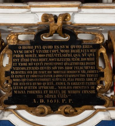 Zdjęcie nr 6: Epitafium uzyskało formę jednokondygnacyjnej, trójosiowej struktury architektonicznej z podstawą i zwieńczeniem, flankowanej po bokach fantazyjnymi uszakami. Część środkową tworzą trzy aedicule rozdzielone dwiema złoconymi kolumnami o korynckich kapitelach i trzonach dekorowanych festonami, a w dolnych partiach pierścieniami. Kolumny podtrzymują pełne belkowanie przechodzące przez całą szerokości kondygnacji, wyłamane nad osiami bocznymi. Same zaś stoją na cokole, który stanowi równocześnie wyłamany gzyms dla podstawy. Pole osi środkowej otrzymało kształt aediculi w formie stojącego prostokąta ze schodkowym zamknięciem u góry, wypełnionym ażurowym, złoconym maswerkiem. Jej wnętrze wypełnia płaskorzeźbione słońce pośród chmur na malowanym tle nieba. Poniżej ukazano panoramę Jerozolimy. Przed aediculą ustawiono na postumencie drewniany krzyż. Na osiach bocznych znalazły się nisze zwieńczone złotymi muszlami oraz złoconą, rytą ornamentyką z motywem czteroliścia. Pierwotnie znajdowały się tu rzeźby Mojżesza i Jana Chrzciciela. Całość po bokach flankują fantazyjne uszaki utworzone ornamentu rollwerkowego oraz z widocznych z profilu półpostaci maurów podtrzymujących belkowanie. Pole uszaków wypełniają pionowo ułożone owalne kartusze o czerwonych polach, stanowiące tło dla dwóch par tarczy herbowych z klejnotami. Z lewej umieszczono herb Dołęga, a z prawej herb Jastrzębiec (bez klejnotu). Po zewnętrznych stronach na belkowaniu ustawiono dwa półsiedzące uskrzydlone anioły (zachował się tylko anioł z prawej strony). Jednocześnie postacie aniołów flankują zwieńczenie epitafium, które tworzy aedicula wypełniona złotą muszlą oraz rytym złoconym ornamentem z motywem czteroliścia. Flankują ją dwa pilastry podtrzymujące wyłamane belkowanie na ich osi. W aediculi ustawiono pełnoplastyczną rzeźbę stojącej kobiety. Została ona ukazana w wydatnym kontrapoście z tułowiem skręconym w lewo, z głową ujętą w trzech czwartych w prawo. Kobieta w podniesionej prawej ręce trzyma srebrny kielich, natomiast w opuszczonej i wspartej o wysuniętą tanecznie lewą nogę otwartą księgę. Postać ma owalną twarz o jasnej karnacji i rumianych policzkach z prostym nosem i wydatnymi ustami. Jej głowę otaczają falujące, brązowe włosy opadające na plecy i  odsłaniające uszy. Ubrana jest w złoconą, mocno drapowaną suknię, która odsłania wysuniętą nogę. Po bokach pilastrów widoczne są dwa uszaki. Na szczycie zwieńczenia dwa pulchne putta prezentują chustę Weroniki, która została równocześnie przytwierdzona do wysokiego impostu wspierającego ażurowy, złocony kwiaton. Na chuście wyobrażono płaskorzeźbioną głowę Chrystusa w cierniowej koronie. Zbawiciel ma owalną twarz z szeroko otwartymi oczami, wydatnym nosem i półotwartymi ustami. Otaczają ją kręcone brązowe włosy oraz podobna, krótka broda. Podstawę epitafium tworzy wykrojona po bokach struktura, w której centrum umieszczono fantazyjnie wyciętą drewnianą, czarną tablicę ze złotym łacińskim napisem &quot;O HOMO FVI QVOD ES SVM QVOD ERIS / NVNC DE(M)VM VIVERE CO(E)PI, MORI DESII, VITA MIHI / PRIOR MORTE(M), MORS PRIA(M) VITA(M) SECV(N)DA(M), QVAE NU(N) QVA(M) / EST I(N)TERI TVRA DEDIT, MO(R)TALITATES  MEOR, HO(C) MIHI / EST ET VXORI MO(S) VENTV(S) POSVI, VIPOST QV(E)ALETHIVERA DVCEDI / NELVSOS, ET CARNIS EXVVIIS SO(L)VTOS  ABHOC PELAG  SECVRA / GLAVSTRA SVB IRE IVABIT, HIC MORTALE DIMIDIVM N(OST)RI, IMMORTA/LE(M) EXPECT ET IM(M)ORTALITATEM, E QVOS VITA CONIVCTOS TENVIT / MORS IDIVISOS CVSTODIRET, SI PIVS ES, PIAS PRECES ADDE, ET / HIC HV(M)O SVB HVMO QVIETA[M], ANIMAE IN COELESTI PATRIA / LETAM  QVIETEM APPRECARE &quot;NICOLAVS CORNITIVS DE / MAGNA POREMBA ET BEATA DE MIROW CONSOR/TES SE(M)PER VAIESL[?]&quot; / A(NNO) D(OMINI) 1611 P(ATRONVS) P(OSVIT)&quot;. Tablicę umieszczono w kartuszu rollwerkowym pomiędzy dwiema konsolami zdobionymi pierwotnie uskrzydlonymi główkami aniołków, podtrzymującymi belkowanie z krzyżem. Poniżej tablicy zastosowano odwrócony łuk odcinkowy z podwieszonym kartuszem rollwerkowym zawieszonym na festonach. W jego wnętrzu przedstawiono tabliczkę z napisem „VITA FUGIT VT HORA”, a po niżej trupią czaszkę ze skrzyżowanymi piszczelami.  
