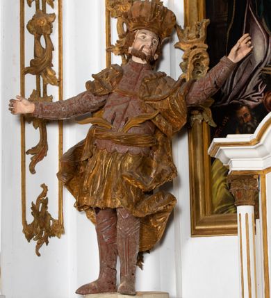 Zdjęcie nr 2: Ustawiona na cokole o nieregularnym planie, pełnoplastyczna rzeźba drążona, ukazuje anonimowego świętego króla. Figura zwrócona jest do widza frontalnie, z głową i torsem wychylonym w lewo i skierowanymi w układzie trzech czwartych. Król stoi w kontrapoście z rękami rozłożonymi na boki. Jest to dojrzały mężczyzna o szczupłej twarzy i podkreślonych kościach policzkowych i linii łuków brwiowych. Charakteryzuje się silnie zadartym, wąskim nosem, pełnymi i lekko rozchylonymi wargami. Jego niewielkie, okrągłe oczy skierowane są w górę. Twarz okala silnie skręcona w dwa pukle broda oraz krótkie, kręcone włosy. Monarcha ubrany jest w tunikę lamowaną frędzlą i sięgającą kolan. Na niej nałożony ma pancerz lekkiej zbroi płytowej z zarękawiami przewiązany w pasie szarfą, za którą ma wetknięte dwie gałązki palmowe. Jego nogi ochraniają nadgolennice i trzewiki. Na ramionach ma narzucony płaszcz podbity gronostajem, zapięty na piersi, którego poły rozwiewają się po bokach postaci. Jego głowę zdobi zamknięta korona o stylizowanych sterczynach. Partie odsłoniętego ciała pokrywa naturalistyczna polichromia, natomiast korona i płaszcz są złocone, a elementy zbroi srebrzone.