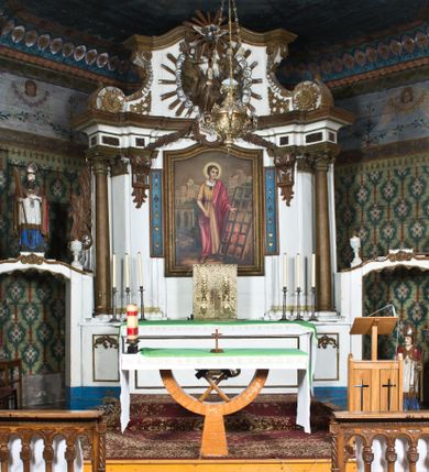Zdjęcie nr 1: Nastawa ołtarzowa na planie półowalu uzyskała architektoniczną, jednokondygnacyjną strukturę ze zwieńczeniem. Jednoosiowe retabulum flankowane bramkami, na których ustawiono figury św. Wojciecha i św. Stanisława. Stipes ołtarz ujmuje drewniana prostopadłościenna obudowa podtrzymująca drewnianą mensą z portatylem. Drewniane antependium ujęte zostało czworoboczną, profilowaną ramą z płaskorzeźbionym i złoconym barankiem apokaliptycznym leżącym na księdze. Na mensie ustawiono współczesne tabernakulum pancerne w formie prostopadłościennej skrzynki. Kondygnacja retabulum ujęta została dwiema parami pilastrów zwróconych delikatnie do wewnątrz oraz silnie wysuniętą do przodu parą kolumn o kapitelach kompozytowych oraz dwie wolutowe konsole. Podpory dźwigają pełne belkowanie wsparte na impostach, o wklęsło-wypukłej linii. Nad polem środkowym widoczne jest połączenie wyłącznie gzymsem przyjmującym formę łuku falistego, wyłamane na osiach podpór. Na środku belkowania umieszczono kartusz, od którego odchodzą dwie girland kwiatowe. Kondygnacje ujmują zredukowane do wici akantowej uszaki. W polu środkowym usytuowano obraz św. Wawrzyńca ujęty profilowaną ramą w formie stojącego prostokąta zamkniętego łukiem nadwieszonym. Zwieńczenie nastawy uzyskało formę leżącego prostokąta flankowanego dwoma spływami wolutowymi opadającymi na belkowanie. Obie woluty dźwigają niepełne, wklęsło-wypukłe belkowanie przyjmujące nad polem formę łuku falistego. W centrum zwieńczenia umieszczono rzeźbioną grupę ze sceną koronacji Marii w srebrzonych obłokach z promieniami. Struktura nastawy jest polichromowana w kolorze białym, kolumny, konsole, profile i ornamentyka pierwotnie były złocone.