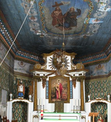 Zdjęcie nr 6: Nastawa ołtarzowa na planie półowalu uzyskała architektoniczną, jednokondygnacyjną strukturę ze zwieńczeniem. Jednoosiowe retabulum flankowane bramkami, na których ustawiono figury św. Wojciecha i św. Stanisława. Stipes ołtarz ujmuje drewniana prostopadłościenna obudowa podtrzymująca drewnianą mensą z portatylem. Drewniane antependium ujęte zostało czworoboczną, profilowaną ramą z płaskorzeźbionym i złoconym barankiem apokaliptycznym leżącym na księdze. Na mensie ustawiono współczesne tabernakulum pancerne w formie prostopadłościennej skrzynki. Kondygnacja retabulum ujęta została dwiema parami pilastrów zwróconych delikatnie do wewnątrz oraz silnie wysuniętą do przodu parą kolumn o kapitelach kompozytowych oraz dwie wolutowe konsole. Podpory dźwigają pełne belkowanie wsparte na impostach, o wklęsło-wypukłej linii. Nad polem środkowym widoczne jest połączenie wyłącznie gzymsem przyjmującym formę łuku falistego, wyłamane na osiach podpór. Na środku belkowania umieszczono kartusz, od którego odchodzą dwie girland kwiatowe. Kondygnacje ujmują zredukowane do wici akantowej uszaki. W polu środkowym usytuowano obraz św. Wawrzyńca ujęty profilowaną ramą w formie stojącego prostokąta zamkniętego łukiem nadwieszonym. Zwieńczenie nastawy uzyskało formę leżącego prostokąta flankowanego dwoma spływami wolutowymi opadającymi na belkowanie. Obie woluty dźwigają niepełne, wklęsło-wypukłe belkowanie przyjmujące nad polem formę łuku falistego. W centrum zwieńczenia umieszczono rzeźbioną grupę ze sceną koronacji Marii w srebrzonych obłokach z promieniami. Struktura nastawy jest polichromowana w kolorze białym, kolumny, konsole, profile i ornamentyka pierwotnie były złocone.