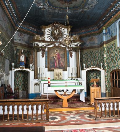 Zdjęcie nr 2: Nastawa ołtarzowa na planie półowalu uzyskała architektoniczną, jednokondygnacyjną strukturę ze zwieńczeniem. Jednoosiowe retabulum flankowane bramkami, na których ustawiono figury św. Wojciecha i św. Stanisława. Stipes ołtarz ujmuje drewniana prostopadłościenna obudowa podtrzymująca drewnianą mensą z portatylem. Drewniane antependium ujęte zostało czworoboczną, profilowaną ramą z płaskorzeźbionym i złoconym barankiem apokaliptycznym leżącym na księdze. Na mensie ustawiono współczesne tabernakulum pancerne w formie prostopadłościennej skrzynki. Kondygnacja retabulum ujęta została dwiema parami pilastrów zwróconych delikatnie do wewnątrz oraz silnie wysuniętą do przodu parą kolumn o kapitelach kompozytowych oraz dwie wolutowe konsole. Podpory dźwigają pełne belkowanie wsparte na impostach, o wklęsło-wypukłej linii. Nad polem środkowym widoczne jest połączenie wyłącznie gzymsem przyjmującym formę łuku falistego, wyłamane na osiach podpór. Na środku belkowania umieszczono kartusz, od którego odchodzą dwie girland kwiatowe. Kondygnacje ujmują zredukowane do wici akantowej uszaki. W polu środkowym usytuowano obraz św. Wawrzyńca ujęty profilowaną ramą w formie stojącego prostokąta zamkniętego łukiem nadwieszonym. Zwieńczenie nastawy uzyskało formę leżącego prostokąta flankowanego dwoma spływami wolutowymi opadającymi na belkowanie. Obie woluty dźwigają niepełne, wklęsło-wypukłe belkowanie przyjmujące nad polem formę łuku falistego. W centrum zwieńczenia umieszczono rzeźbioną grupę ze sceną koronacji Marii w srebrzonych obłokach z promieniami. Struktura nastawy jest polichromowana w kolorze białym, kolumny, konsole, profile i ornamentyka pierwotnie były złocone.