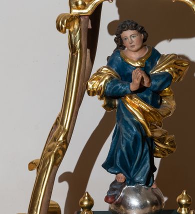 Zdjęcie nr 1: Feretron w formie pełnoplastycznej figury Matki Boskiej Niepokalanie Poczętej. Rzeźba usytuowana jest na prostokątnej podstawie o wklęsłym bokach oraz nakryta baldachimem wspartym na czterech wolutach, zwieńczonym poduszką, na której leży korona w typie zamkniętym. Zwrócona frontalnie sylwetka Matki Boskiej stojącej w kontrapoście na globie osadzona jest na profilowanym cokole. Maria prawą stopą depcze głowę węża z jabłkiem w pysku. Jej dłonie złożone są w geście modlitwy, głowa przechylona na prawe ramię. Pociągłą twarz o pełnych policzkach, płytko rzeźbionych rysach okalają krótkie, silnie skręcone pukle. Maria jest ubrana w niebieską suknię oraz przerzucony przez przedramiona płaszcz spiralnie oplatający jej sylwetkę. Tkanina jest  rozwiana i dynamicznie drapowana.