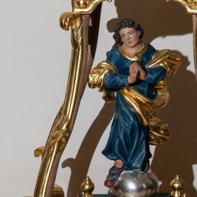 Zdjęcie nr 1: Feretron w formie pełnoplastycznej figury Matki Boskiej Niepokalanie Poczętej. Rzeźba usytuowana jest na prostokątnej podstawie o wklęsłym bokach oraz nakryta baldachimem wspartym na czterech wolutach, zwieńczonym poduszką, na której leży korona w typie zamkniętym. Zwrócona frontalnie sylwetka Matki Boskiej stojącej w kontrapoście na globie osadzona jest na profilowanym cokole. Maria prawą stopą depcze głowę węża z jabłkiem w pysku. Jej dłonie złożone są w geście modlitwy, głowa przechylona na prawe ramię. Pociągłą twarz o pełnych policzkach, płytko rzeźbionych rysach okalają krótkie, silnie skręcone pukle. Maria jest ubrana w niebieską suknię oraz przerzucony przez przedramiona płaszcz spiralnie oplatający jej sylwetkę. Tkanina jest  rozwiana i dynamicznie drapowana.