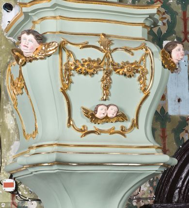 Zdjęcie nr 5: Koszowa ambona przyścienna z baldachimem uzyskała formę architektoniczną. Czworoboczny kosz otrzymał ścięte naroża. Jego boki mają linię wklęsło-wypukłą. Wypełniono je pseudopłycinami utworzonymi ze złoconych ceownic, tworzących kartusze z festonami i girlandami. W polu środkowym oraz w narożach kosza ambony umieszczono uskrzydlone główki anielskie. Całość kosza została zwieńczona profilowanym gzymsem. U dołu podwieszono ażurową, złoconą strukturę utworzoną z czterech wolut i kwiatonu. W zaplecku ambony usytuowano dwuskrzydłowe drzwi ujęte pilastrami o gładkich trzonach. Górne części pilastrów udekorowano podwieszonymi festonami. Baldachim uzyskał rzut prostokąta o ściętych narożach oraz wklęsło-wypukłe boki. Został wsparty na dwóch konsolach, na których usytuowano pełne belkowanie z podwieszonym lambrekinem. Partię fryzu zdobi złocony ornament roślinnym, uzupełniony kwiatami. Całość baldachimu zwieńczono płaskorzeźbioną, złoconą i srebrzoną gołębicą Ducha Świętego w srebrzonych obłokach na tle glorii promienistej. Strukturę ambony pomalowano w kolorze jasnozielonym, natomiast partie ornamentów zostały wyzłocone, zaś karnacje uskrzydlonych aniołków polichromowano.