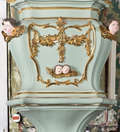 Zdjęcie nr 4: Koszowa ambona przyścienna z baldachimem uzyskała formę architektoniczną. Czworoboczny kosz otrzymał ścięte naroża. Jego boki mają linię wklęsło-wypukłą. Wypełniono je pseudopłycinami utworzonymi ze złoconych ceownic, tworzących kartusze z festonami i girlandami. W polu środkowym oraz w narożach kosza ambony umieszczono uskrzydlone główki anielskie. Całość kosza została zwieńczona profilowanym gzymsem. U dołu podwieszono ażurową, złoconą strukturę utworzoną z czterech wolut i kwiatonu. W zaplecku ambony usytuowano dwuskrzydłowe drzwi ujęte pilastrami o gładkich trzonach. Górne części pilastrów udekorowano podwieszonymi festonami. Baldachim uzyskał rzut prostokąta o ściętych narożach oraz wklęsło-wypukłe boki. Został wsparty na dwóch konsolach, na których usytuowano pełne belkowanie z podwieszonym lambrekinem. Partię fryzu zdobi złocony ornament roślinnym, uzupełniony kwiatami. Całość baldachimu zwieńczono płaskorzeźbioną, złoconą i srebrzoną gołębicą Ducha Świętego w srebrzonych obłokach na tle glorii promienistej. Strukturę ambony pomalowano w kolorze jasnozielonym, natomiast partie ornamentów zostały wyzłocone, zaś karnacje uskrzydlonych aniołków polichromowano.