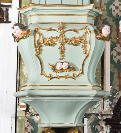 Zdjęcie nr 1: Koszowa ambona przyścienna z baldachimem uzyskała formę architektoniczną. Czworoboczny kosz otrzymał ścięte naroża. Jego boki mają linię wklęsło-wypukłą. Wypełniono je pseudopłycinami utworzonymi ze złoconych ceownic, tworzących kartusze z festonami i girlandami. W polu środkowym oraz w narożach kosza ambony umieszczono uskrzydlone główki anielskie. Całość kosza została zwieńczona profilowanym gzymsem. U dołu podwieszono ażurową, złoconą strukturę utworzoną z czterech wolut i kwiatonu. W zaplecku ambony usytuowano dwuskrzydłowe drzwi ujęte pilastrami o gładkich trzonach. Górne części pilastrów udekorowano podwieszonymi festonami. Baldachim uzyskał rzut prostokąta o ściętych narożach oraz wklęsło-wypukłe boki. Został wsparty na dwóch konsolach, na których usytuowano pełne belkowanie z podwieszonym lambrekinem. Partię fryzu zdobi złocony ornament roślinnym, uzupełniony kwiatami. Całość baldachimu zwieńczono płaskorzeźbioną, złoconą i srebrzoną gołębicą Ducha Świętego w srebrzonych obłokach na tle glorii promienistej. Strukturę ambony pomalowano w kolorze jasnozielonym, natomiast partie ornamentów zostały wyzłocone, zaś karnacje uskrzydlonych aniołków polichromowano.