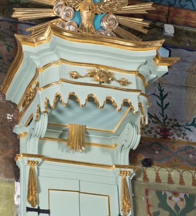 Zdjęcie nr 3: Koszowa ambona przyścienna z baldachimem uzyskała formę architektoniczną. Czworoboczny kosz otrzymał ścięte naroża. Jego boki mają linię wklęsło-wypukłą. Wypełniono je pseudopłycinami utworzonymi ze złoconych ceownic, tworzących kartusze z festonami i girlandami. W polu środkowym oraz w narożach kosza ambony umieszczono uskrzydlone główki anielskie. Całość kosza została zwieńczona profilowanym gzymsem. U dołu podwieszono ażurową, złoconą strukturę utworzoną z czterech wolut i kwiatonu. W zaplecku ambony usytuowano dwuskrzydłowe drzwi ujęte pilastrami o gładkich trzonach. Górne części pilastrów udekorowano podwieszonymi festonami. Baldachim uzyskał rzut prostokąta o ściętych narożach oraz wklęsło-wypukłe boki. Został wsparty na dwóch konsolach, na których usytuowano pełne belkowanie z podwieszonym lambrekinem. Partię fryzu zdobi złocony ornament roślinnym, uzupełniony kwiatami. Całość baldachimu zwieńczono płaskorzeźbioną, złoconą i srebrzoną gołębicą Ducha Świętego w srebrzonych obłokach na tle glorii promienistej. Strukturę ambony pomalowano w kolorze jasnozielonym, natomiast partie ornamentów zostały wyzłocone, zaś karnacje uskrzydlonych aniołków polichromowano.