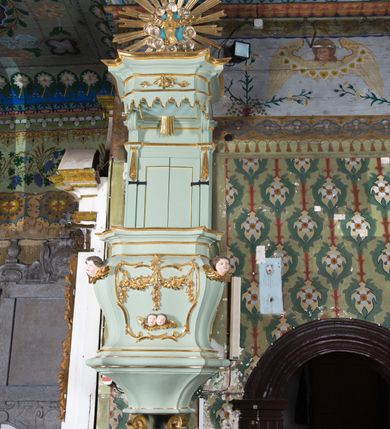 Zdjęcie nr 2: Koszowa ambona przyścienna z baldachimem uzyskała formę architektoniczną. Czworoboczny kosz otrzymał ścięte naroża. Jego boki mają linię wklęsło-wypukłą. Wypełniono je pseudopłycinami utworzonymi ze złoconych ceownic, tworzących kartusze z festonami i girlandami. W polu środkowym oraz w narożach kosza ambony umieszczono uskrzydlone główki anielskie. Całość kosza została zwieńczona profilowanym gzymsem. U dołu podwieszono ażurową, złoconą strukturę utworzoną z czterech wolut i kwiatonu. W zaplecku ambony usytuowano dwuskrzydłowe drzwi ujęte pilastrami o gładkich trzonach. Górne części pilastrów udekorowano podwieszonymi festonami. Baldachim uzyskał rzut prostokąta o ściętych narożach oraz wklęsło-wypukłe boki. Został wsparty na dwóch konsolach, na których usytuowano pełne belkowanie z podwieszonym lambrekinem. Partię fryzu zdobi złocony ornament roślinnym, uzupełniony kwiatami. Całość baldachimu zwieńczono płaskorzeźbioną, złoconą i srebrzoną gołębicą Ducha Świętego w srebrzonych obłokach na tle glorii promienistej. Strukturę ambony pomalowano w kolorze jasnozielonym, natomiast partie ornamentów zostały wyzłocone, zaś karnacje uskrzydlonych aniołków polichromowano.