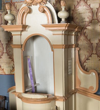 Zdjęcie nr 1: Drewniany konfesjonał uzyskał rozbudowaną formę architektoniczną, ustawioną na niewysokim cokole na rzucie prostokąta zamkniętego łukiem nadwieszonym. Przedpiersie ma wypukły kształt z jednoskrzydłowymi drzwiczkami zwieńczonymi profilowanym gzymsem. Całą strukturę ujęto pilastrami o gładkich trzonach dźwigającymi odcinki pełnego belkowania. Na ich osiach ustawiono płomieniste wazony. W zwieńczeniu znalazły się dwie, przeciwstawnie ustawione woluty oraz muszla palmetowa na tle skrzyżowanych gałązek laurowych. Zaplecek uzyskał gładką formę pozbawioną dekoracji. Całość ujęto po bokach spływami wolutowymi z ustawionymi na ich szczycie wazonikami (brak prawego wazonika). Strukturę konfesjonału pomalowano farbą olejną w kolorze jasnożółtym, zaś detale architektoniczne w kolorze pomarańczowym.