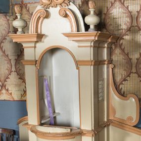 Zdjęcie nr 1: Drewniany konfesjonał uzyskał rozbudowaną formę architektoniczną, ustawioną na niewysokim cokole na rzucie prostokąta zamkniętego łukiem nadwieszonym. Przedpiersie ma wypukły kształt z jednoskrzydłowymi drzwiczkami zwieńczonymi profilowanym gzymsem. Całą strukturę ujęto pilastrami o gładkich trzonach dźwigającymi odcinki pełnego belkowania. Na ich osiach ustawiono płomieniste wazony. W zwieńczeniu znalazły się dwie, przeciwstawnie ustawione woluty oraz muszla palmetowa na tle skrzyżowanych gałązek laurowych. Zaplecek uzyskał gładką formę pozbawioną dekoracji. Całość ujęto po bokach spływami wolutowymi z ustawionymi na ich szczycie wazonikami (brak prawego wazonika). Strukturę konfesjonału pomalowano farbą olejną w kolorze jasnożółtym, zaś detale architektoniczne w kolorze pomarańczowym.