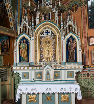 Zdjęcie nr 3: Obraz uzyskał kształt wydłużonego, stojącego prostokąta. W centrum kompozycji wyobrażono w całej postaci ukazaną frontalne Matkę Boską. Madonna została ujęta w kontrapoście, z nieznacznie ugiętymi w kolanach nogami, stojąc na półksiężycu opartym na globie ziemskim, wokół którego owinięty jest wąż z jabłkiem w paszczy. Marię ukazano jako młodą kobietę z rozsuniętymi rękami na wysokości bioder. W prawej ręce trzyma dwukwiatową lilię na długiej łodydze. Lilię przykrywa srebrzona i złocona drewniana sukienka. Niepokalana ma owalną, pełną twarz o rumianych policzkach, mięsistym nosie i ustach. Oczy ma skierowane ku górze. Na głowie widać długie, brązowe włosy spływające na ramiona. Głowę otacza promienisty, płaskorzeźbiony i złocony nimb oraz tak samo wykonane 12 gwiazd. Postać Matki Boskiej, podobnie, jak i glob z wężem, przykrywa złocona, drewniana sukienka, która zapewne odzwierciedla pierwotny układ szat Madonny. Maria ubrana jest w długi płaszcz spięty romboidalną broszą pod szyją, którego poły opadając ku dołowi odsłaniają spodnią sukienkę spiętą w talii paskiem z owalną sprzączką. Płaszcz, podbity gładką podszewką i suknię dekorują krótkie gałązki róż. Ponad Niepokalaną wyobrażono gołębicę Ducha Świętego z rozpostartymi skrzydłami na tle obłoków, także okrytą drewnianą, płaskorzeźbioną i złoconą sukienką. Od głowy gołębicy wychodzi wiązka promieni skierowana w stronę Marii. Postać Madonny otaczają obłoki usytuowane przy krawędziach obrazu, na tle których ukazano po cztery uskrzydlone główki aniołków ułożone wertykalnie. Zarówno obłoki, jak i skrzydła przykrywa drewniana sukienka, srebrzona w partii obłoków, a złocona w części przykrywającej skrzydła. Drewniana sukienka zdobiąca obraz odsłania tylko partię twarzy namalowanych postaci oraz dłonie i stopy.