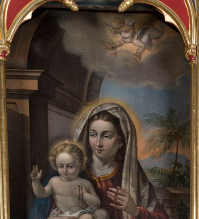 Zdjęcie nr 2: Obraz uzyskał kształt stojącego prostokąta. W centrum kompozycji wyobrażono w półpostaci siedzącą Matkę Boską z Dzieciątkiem na tle szerokiej arkady, której górna część ginie w obłokach. Madonna zwrócona w trzech czwartych w prawo prawą dłonią podtrzymuje siedzące na jej kolanach Dzieciątko Jezus. Natomiast w uniesionej w jego stronę lewej ręce trzyma czerwony różaniec przewieszony przez dłoń. Maria ma owalną twarz o rumianych policzkach, wydatny prosty nos i wąskie usta. Spojrzenie jej migdałowych oczku skierowane jest w stronę Dzieciątka. Głowę Marii otaczają jasnobrązowe, falujące włosy spływające na ramiona. Madonna ubrana jest w szkarłatną suknię z dużym dekoltem odsłaniającym jej szyję oraz rąbek spodniej, białej koszuli. Suknia pod szyją lamowana jest złocistą taśmą. Natomiast bufiaste rękawy przy dłoniach spinają wąskie mankiety. Jej ramiona okrywa niebieski płaszcz przykrywający także jej kolana. Na głowie widoczna jest jasnobeżowa chusta dekorowana poprzecznymi błękitno-czerwonymi, delikatnymi paseczkami, która okrywa także ramiona. Trzymane przez nią Dzieciątko siedzi na bladoczerwonej poduszce zdobionej złocistym obszyciem i chwostami wiszącymi przy jej rogach. Mały Jezus ujęty został w trzech czwartych w lewo. Prawą rękę unosi w geście błogosławieństwa, w lewej zaś przyciśniętej do piersi trzyma czerwony różaniec. Nóżki ma skrzyżowane na poduszce. Jego nagie ciało okrywa tylko w partii bioder biała pieluszka. Dzieciątko ma owalną twarz otoczoną jasnymi, falującymi włosami. Charakteryzują ją wysokie czoło, drobny nos i usta oraz wyraziste spojrzenie skierowane na widza. Głowę Jezusa, podobnie jak i Marii otacza nieznaczna poświata. Ponad Madonną z Dzieciątkiem widoczny jest na tle rozjaśnionych obłoków mały, uskrzydlony, nagi aniołek o jasnych włosach, niosący w rękach gałązki różane. Towarzyszy mu uskrzydlona, pucułowata główka innego aniołka. Za Marią widoczony jest fragment balustrady, za którą znajduje się egzotyczna roślinność z palmą daktylową w tle.