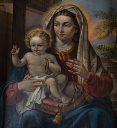 Zdjęcie nr 1: Obraz uzyskał kształt stojącego prostokąta. W centrum kompozycji wyobrażono w półpostaci siedzącą Matkę Boską z Dzieciątkiem na tle szerokiej arkady, której górna część ginie w obłokach. Madonna zwrócona w trzech czwartych w prawo prawą dłonią podtrzymuje siedzące na jej kolanach Dzieciątko Jezus. Natomiast w uniesionej w jego stronę lewej ręce trzyma czerwony różaniec przewieszony przez dłoń. Maria ma owalną twarz o rumianych policzkach, wydatny prosty nos i wąskie usta. Spojrzenie jej migdałowych oczku skierowane jest w stronę Dzieciątka. Głowę Marii otaczają jasnobrązowe, falujące włosy spływające na ramiona. Madonna ubrana jest w szkarłatną suknię z dużym dekoltem odsłaniającym jej szyję oraz rąbek spodniej, białej koszuli. Suknia pod szyją lamowana jest złocistą taśmą. Natomiast bufiaste rękawy przy dłoniach spinają wąskie mankiety. Jej ramiona okrywa niebieski płaszcz przykrywający także jej kolana. Na głowie widoczna jest jasnobeżowa chusta dekorowana poprzecznymi błękitno-czerwonymi, delikatnymi paseczkami, która okrywa także ramiona. Trzymane przez nią Dzieciątko siedzi na bladoczerwonej poduszce zdobionej złocistym obszyciem i chwostami wiszącymi przy jej rogach. Mały Jezus ujęty został w trzech czwartych w lewo. Prawą rękę unosi w geście błogosławieństwa, w lewej zaś przyciśniętej do piersi trzyma czerwony różaniec. Nóżki ma skrzyżowane na poduszce. Jego nagie ciało okrywa tylko w partii bioder biała pieluszka. Dzieciątko ma owalną twarz otoczoną jasnymi, falującymi włosami. Charakteryzują ją wysokie czoło, drobny nos i usta oraz wyraziste spojrzenie skierowane na widza. Głowę Jezusa, podobnie jak i Marii otacza nieznaczna poświata. Ponad Madonną z Dzieciątkiem widoczny jest na tle rozjaśnionych obłoków mały, uskrzydlony, nagi aniołek o jasnych włosach, niosący w rękach gałązki różane. Towarzyszy mu uskrzydlona, pucułowata główka innego aniołka. Za Marią widoczony jest fragment balustrady, za którą znajduje się egzotyczna roślinność z palmą daktylową w tle.