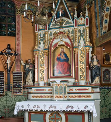 Zdjęcie nr 12: Ołtarz Najświętszego Serca Pana Jezusa uzyskał formę drewnianej i polichromowanej przyściennej struktury architektonicznej. Jest to założenie jednoosiowe i jednokondygnacyjne ze zwieńczeniem. Drewniana mensa wspiera się na prostopadłościennym drewnianym stipesie ołtarzowym flankowanym po bokach kolumienkami na wysokich cokolikach. Antepedium zostało podzielone obramionymi podłużnymi dwiema płycinami. Ich wewnętrzne krótsze boki są wklęsłe. W centrum pomiędzy płycinami znajduje się złocony czteroliść w owalu z wpisanym w środku krzyżem równoramiennym. Nastawa została ustawiona na zdwojonym cokole z przylegającym do niego na środku wielobocznym tabernakulum. Prostopadłościenne tabernakulum o ściętych narożach dekorowane jest płycinami ozdobionymi przenikającymi się ostrołukami z noskami. Drzwiczki zdobią litery alfa i omega. Górną część tabernakulum zamyka profilowany gzyms, natomiast dolną dekoruje pseudogotycki fryz z motywem czteroliścia wpisanego w romboidalną kratownicę. Podobny fryz znajduje się w dolnej cześć cokołu nastawy. Z kolei górną zdobią podłużne płyciny ze złoconą dekoracją maswerkową. Z cokołu wyrasta architektoniczna struktura utworzona z centralnej aediculi flankowanej parami kolumn o korynckich, złoconych kapitelach i czerwonych trzonach dekorowanych złotym ornamentem geometrycznym. Kolumny zostały ustawione na niewielkich postumentach. Podtrzymują one imposty z cokolikami z rozetami, z których wyrastają złocone, skręcone mniejsze kolumienki z fantazyjnymi kapitelami podtrzymującymi czworoboczne sterczyny przechodzące w pinakle zwieńczone kwiatonami. W dolnej części pomiędzy kolumnami zastosowano wydłużone płyciny zwieńczone trójbocznie z wpisanymi ostrołukami, wypełnionymi złoconym ornamentem astwerkowym. Natomiast w górnej części pomiędzy skręconymi kolumienkami oraz w tle arkady wieńczącej aediculę zastosowano płyciny dekorowane złoconymi ostrołukami z noskami. Ponad bocznymi płycinami wypiętrzono po bokach dwa mniejsze trójkątne szczyty zakończone pinaklami. Większy trójkątny szczyt, wypełniony obrazem z przedstawieniem Niepokalanego Serca Najświętszej Marii Panny, wznosi się nad centralną aediculą zakończoną ostrołukowo. W górnej części ostrołuk aediculi wypełnia ażurowy fantazyjny maswerk. W aediculi znajduje się obraz Najświętszego Serca Pana Jezusa, a na zasuwach obraz Matki Boskiej Różańcowej i św. Barbary. Nastawę po bokach flankują pełnoplastyczne figury św. Dominika oraz św. Katarzyny Sieneńskiej. Całość polichromowana jest w odcieniach seledynowej zieleni, rozbielonej czerwieni oraz błękintawej szarości. Partie ornamentów i profile płycin są złocone.