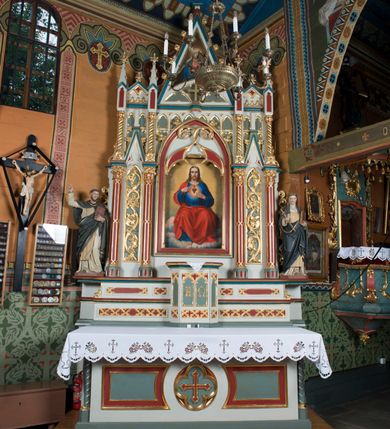 Zdjęcie nr 2: Ołtarz Najświętszego Serca Pana Jezusa uzyskał formę drewnianej i polichromowanej przyściennej struktury architektonicznej. Jest to założenie jednoosiowe i jednokondygnacyjne ze zwieńczeniem. Drewniana mensa wspiera się na prostopadłościennym drewnianym stipesie ołtarzowym flankowanym po bokach kolumienkami na wysokich cokolikach. Antepedium zostało podzielone obramionymi podłużnymi dwiema płycinami. Ich wewnętrzne krótsze boki są wklęsłe. W centrum pomiędzy płycinami znajduje się złocony czteroliść w owalu z wpisanym w środku krzyżem równoramiennym. Nastawa została ustawiona na zdwojonym cokole z przylegającym do niego na środku wielobocznym tabernakulum. Prostopadłościenne tabernakulum o ściętych narożach dekorowane jest płycinami ozdobionymi przenikającymi się ostrołukami z noskami. Drzwiczki zdobią litery alfa i omega. Górną część tabernakulum zamyka profilowany gzyms, natomiast dolną dekoruje pseudogotycki fryz z motywem czteroliścia wpisanego w romboidalną kratownicę. Podobny fryz znajduje się w dolnej cześć cokołu nastawy. Z kolei górną zdobią podłużne płyciny ze złoconą dekoracją maswerkową. Z cokołu wyrasta architektoniczna struktura utworzona z centralnej aediculi flankowanej parami kolumn o korynckich, złoconych kapitelach i czerwonych trzonach dekorowanych złotym ornamentem geometrycznym. Kolumny zostały ustawione na niewielkich postumentach. Podtrzymują one imposty z cokolikami z rozetami, z których wyrastają złocone, skręcone mniejsze kolumienki z fantazyjnymi kapitelami podtrzymującymi czworoboczne sterczyny przechodzące w pinakle zwieńczone kwiatonami. W dolnej części pomiędzy kolumnami zastosowano wydłużone płyciny zwieńczone trójbocznie z wpisanymi ostrołukami, wypełnionymi złoconym ornamentem astwerkowym. Natomiast w górnej części pomiędzy skręconymi kolumienkami oraz w tle arkady wieńczącej aediculę zastosowano płyciny dekorowane złoconymi ostrołukami z noskami. Ponad bocznymi płycinami wypiętrzono po bokach dwa mniejsze trójkątne szczyty zakończone pinaklami. Większy trójkątny szczyt, wypełniony obrazem z przedstawieniem Niepokalanego Serca Najświętszej Marii Panny, wznosi się nad centralną aediculą zakończoną ostrołukowo. W górnej części ostrołuk aediculi wypełnia ażurowy fantazyjny maswerk. W aediculi znajduje się obraz Najświętszego Serca Pana Jezusa, a na zasuwach obraz Matki Boskiej Różańcowej i św. Barbary. Nastawę po bokach flankują pełnoplastyczne figury św. Dominika oraz św. Katarzyny Sieneńskiej. Całość polichromowana jest w odcieniach seledynowej zieleni, rozbielonej czerwieni oraz błękintawej szarości. Partie ornamentów i profile płycin są złocone.