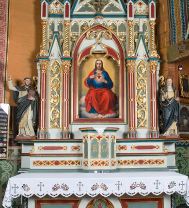 Zdjęcie nr 1: Ołtarz Najświętszego Serca Pana Jezusa uzyskał formę drewnianej i polichromowanej przyściennej struktury architektonicznej. Jest to założenie jednoosiowe i jednokondygnacyjne ze zwieńczeniem. Drewniana mensa wspiera się na prostopadłościennym drewnianym stipesie ołtarzowym flankowanym po bokach kolumienkami na wysokich cokolikach. Antepedium zostało podzielone obramionymi podłużnymi dwiema płycinami. Ich wewnętrzne krótsze boki są wklęsłe. W centrum pomiędzy płycinami znajduje się złocony czteroliść w owalu z wpisanym w środku krzyżem równoramiennym. Nastawa została ustawiona na zdwojonym cokole z przylegającym do niego na środku wielobocznym tabernakulum. Prostopadłościenne tabernakulum o ściętych narożach dekorowane jest płycinami ozdobionymi przenikającymi się ostrołukami z noskami. Drzwiczki zdobią litery alfa i omega. Górną część tabernakulum zamyka profilowany gzyms, natomiast dolną dekoruje pseudogotycki fryz z motywem czteroliścia wpisanego w romboidalną kratownicę. Podobny fryz znajduje się w dolnej cześć cokołu nastawy. Z kolei górną zdobią podłużne płyciny ze złoconą dekoracją maswerkową. Z cokołu wyrasta architektoniczna struktura utworzona z centralnej aediculi flankowanej parami kolumn o korynckich, złoconych kapitelach i czerwonych trzonach dekorowanych złotym ornamentem geometrycznym. Kolumny zostały ustawione na niewielkich postumentach. Podtrzymują one imposty z cokolikami z rozetami, z których wyrastają złocone, skręcone mniejsze kolumienki z fantazyjnymi kapitelami podtrzymującymi czworoboczne sterczyny przechodzące w pinakle zwieńczone kwiatonami. W dolnej części pomiędzy kolumnami zastosowano wydłużone płyciny zwieńczone trójbocznie z wpisanymi ostrołukami, wypełnionymi złoconym ornamentem astwerkowym. Natomiast w górnej części pomiędzy skręconymi kolumienkami oraz w tle arkady wieńczącej aediculę zastosowano płyciny dekorowane złoconymi ostrołukami z noskami. Ponad bocznymi płycinami wypiętrzono po bokach dwa mniejsze trójkątne szczyty zakończone pinaklami. Większy trójkątny szczyt, wypełniony obrazem z przedstawieniem Niepokalanego Serca Najświętszej Marii Panny, wznosi się nad centralną aediculą zakończoną ostrołukowo. W górnej części ostrołuk aediculi wypełnia ażurowy fantazyjny maswerk. W aediculi znajduje się obraz Najświętszego Serca Pana Jezusa, a na zasuwach obraz Matki Boskiej Różańcowej i św. Barbary. Nastawę po bokach flankują pełnoplastyczne figury św. Dominika oraz św. Katarzyny Sieneńskiej. Całość polichromowana jest w odcieniach seledynowej zieleni, rozbielonej czerwieni oraz błękintawej szarości. Partie ornamentów i profile płycin są złocone.