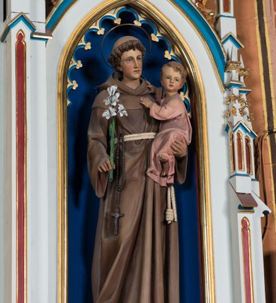 Zdjęcie nr 2: Gipsowa, pełnoplastyczna figura św. Antoniego z Dzieciątkiem Jezus ustawiona została na ciemnym, niewysokim prostopadłościennym postumencie o ściętych narożach. Święty został ukazany całopostaciowo w lekkim kontrapoście w nieznacznym esowatym wygięciu. W prawej ręce trzyma lilię, a na lewym ramieniu małego Jezusa. Antoniego przedstawiono jako młodego mężczyznę o kędzierzawych włosach z tonsurą. Ma pociągłą, lekko wychudzoną twarz o jasnej karnacji z wyrazistymi oczami, prostym nosem i wydatnymi ustami. Głowę otacza owalny druciany nimb. Ubrany jest w brązowy habit franciszkański przewiązany podwójnym sznurem z węzłem przy jednym z końców. Z prawej strony przez sznur przewieszony jest czarny różaniec. Trzymane przez niego Dzieciątko zostało ujęte w trzech czwartych w prawo. Mały Jezus obiema rączkami zdaje się trzymać Antoniego. Głowę o jasnych włosach Zbawiciel odwraca w stronę widza, patrząc przed siebie. Ma wysokie czoło, rumiane policzki, wyraziste oczy i usta oraz prosty nos. Ubrany jest w bladoróżową tunikę, odsłaniającą fragment lewej stopy.