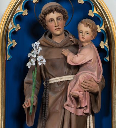 Zdjęcie nr 1: Gipsowa, pełnoplastyczna figura św. Antoniego z Dzieciątkiem Jezus ustawiona została na ciemnym, niewysokim prostopadłościennym postumencie o ściętych narożach. Święty został ukazany całopostaciowo w lekkim kontrapoście w nieznacznym esowatym wygięciu. W prawej ręce trzyma lilię, a na lewym ramieniu małego Jezusa. Antoniego przedstawiono jako młodego mężczyznę o kędzierzawych włosach z tonsurą. Ma pociągłą, lekko wychudzoną twarz o jasnej karnacji z wyrazistymi oczami, prostym nosem i wydatnymi ustami. Głowę otacza owalny druciany nimb. Ubrany jest w brązowy habit franciszkański przewiązany podwójnym sznurem z węzłem przy jednym z końców. Z prawej strony przez sznur przewieszony jest czarny różaniec. Trzymane przez niego Dzieciątko zostało ujęte w trzech czwartych w prawo. Mały Jezus obiema rączkami zdaje się trzymać Antoniego. Głowę o jasnych włosach Zbawiciel odwraca w stronę widza, patrząc przed siebie. Ma wysokie czoło, rumiane policzki, wyraziste oczy i usta oraz prosty nos. Ubrany jest w bladoróżową tunikę, odsłaniającą fragment lewej stopy.
