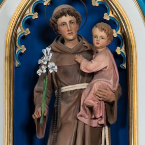 Zdjęcie nr 1: Gipsowa, pełnoplastyczna figura św. Antoniego z Dzieciątkiem Jezus ustawiona została na ciemnym, niewysokim prostopadłościennym postumencie o ściętych narożach. Święty został ukazany całopostaciowo w lekkim kontrapoście w nieznacznym esowatym wygięciu. W prawej ręce trzyma lilię, a na lewym ramieniu małego Jezusa. Antoniego przedstawiono jako młodego mężczyznę o kędzierzawych włosach z tonsurą. Ma pociągłą, lekko wychudzoną twarz o jasnej karnacji z wyrazistymi oczami, prostym nosem i wydatnymi ustami. Głowę otacza owalny druciany nimb. Ubrany jest w brązowy habit franciszkański przewiązany podwójnym sznurem z węzłem przy jednym z końców. Z prawej strony przez sznur przewieszony jest czarny różaniec. Trzymane przez niego Dzieciątko zostało ujęte w trzech czwartych w prawo. Mały Jezus obiema rączkami zdaje się trzymać Antoniego. Głowę o jasnych włosach Zbawiciel odwraca w stronę widza, patrząc przed siebie. Ma wysokie czoło, rumiane policzki, wyraziste oczy i usta oraz prosty nos. Ubrany jest w bladoróżową tunikę, odsłaniającą fragment lewej stopy.
