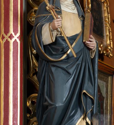 Zdjęcie nr 2: Pełnoplastyczna figura stojącej św. Katarzyny Sieneńskiej ustawiona została na niskim prostopadłościennym postumencie o ściętych narożach. Święta została przedstawiona w lekkim kontrapoście jako młoda kobieta. W uniesionej prawej ręce trzyma złocony krucyfiks, a  w lewej przyciśniętej do piersi zamknietą księgę. Ma owalną, pełną twarz o jasnej karnacji. Charakteryzują ją małe usta, prosty nos oraz wydatny podbródek. Katarzyna ubrana jest w biały habit dominikański z kremowym szkaplerzem, lamowanym złotą taśmą. Na ramionach ma narzucony ciemnoszary płaszcz lamowany przy krawędziach złotą taśmą. Na głowie ma zakonny kornet z podwiką i szaroczarny welon oraz złoconą koronę cierniową. 