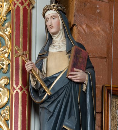 Zdjęcie nr 1: Pełnoplastyczna figura stojącej św. Katarzyny Sieneńskiej ustawiona została na niskim prostopadłościennym postumencie o ściętych narożach. Święta została przedstawiona w lekkim kontrapoście jako młoda kobieta. W uniesionej prawej ręce trzyma złocony krucyfiks, a  w lewej przyciśniętej do piersi zamknietą księgę. Ma owalną, pełną twarz o jasnej karnacji. Charakteryzują ją małe usta, prosty nos oraz wydatny podbródek. Katarzyna ubrana jest w biały habit dominikański z kremowym szkaplerzem, lamowanym złotą taśmą. Na ramionach ma narzucony ciemnoszary płaszcz lamowany przy krawędziach złotą taśmą. Na głowie ma zakonny kornet z podwiką i szaroczarny welon oraz złoconą koronę cierniową. 