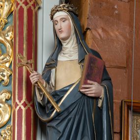 Zdjęcie nr 1: Pełnoplastyczna figura stojącej św. Katarzyny Sieneńskiej ustawiona została na niskim prostopadłościennym postumencie o ściętych narożach. Święta została przedstawiona w lekkim kontrapoście jako młoda kobieta. W uniesionej prawej ręce trzyma złocony krucyfiks, a  w lewej przyciśniętej do piersi zamknietą księgę. Ma owalną, pełną twarz o jasnej karnacji. Charakteryzują ją małe usta, prosty nos oraz wydatny podbródek. Katarzyna ubrana jest w biały habit dominikański z kremowym szkaplerzem, lamowanym złotą taśmą. Na ramionach ma narzucony ciemnoszary płaszcz lamowany przy krawędziach złotą taśmą. Na głowie ma zakonny kornet z podwiką i szaroczarny welon oraz złoconą koronę cierniową. 