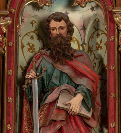 Zdjęcie nr 1: Pełnoplastyczna, polichromowana figura ustawiona na niewysokim, prostopadłościennym postumencie ukazuje św. Pawła Apostoła. Święty został przedstawiony frontalnie w nieznacznym kontrapoście jako dojrzały, ciemnowłosy mężczyzna z okazałą, rozwianą brodą. W lewej dłoni podtrzymuje otwartą księgę, a prawą opiera na mieczu zwróconym ostrzem ku ziemi. Jednocześnie lekko pochyla głowę do przodu. Apostoł ma pociągłą twarz o jasnej karnacji, wysokie czoło, prosty nos i zamyślone spojrzenie. Ubrany jest w długą ciemnoniebieską tunikę spiętą w pasie oraz obszerny, czerwony płaszcz ze złotą lamówką, spięty na prawym ramieniu fibulą. Na cokole widnieje złocona inskrypcja „Św(ięty) Paweł".