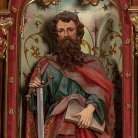 Zdjęcie nr 1: Pełnoplastyczna, polichromowana figura ustawiona na niewysokim, prostopadłościennym postumencie ukazuje św. Pawła Apostoła. Święty został przedstawiony frontalnie w nieznacznym kontrapoście jako dojrzały, ciemnowłosy mężczyzna z okazałą, rozwianą brodą. W lewej dłoni podtrzymuje otwartą księgę, a prawą opiera na mieczu zwróconym ostrzem ku ziemi. Jednocześnie lekko pochyla głowę do przodu. Apostoł ma pociągłą twarz o jasnej karnacji, wysokie czoło, prosty nos i zamyślone spojrzenie. Ubrany jest w długą ciemnoniebieską tunikę spiętą w pasie oraz obszerny, czerwony płaszcz ze złotą lamówką, spięty na prawym ramieniu fibulą. Na cokole widnieje złocona inskrypcja „Św(ięty) Paweł".