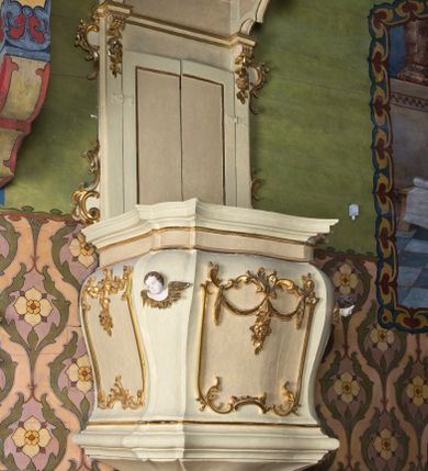 Zdjęcie nr 1: Ambona wisząca, przyścienna z baldachimem. Kosz czworoboczny o ściętych narożach, lekko wybrzuszony w górnej części. W polach płyciny utworzone z ornamentu rocaille oraz podwieszone festony roślinne; w narożach polichromowane i złocone uskrzydlone główki anielskie. U dołu kosza ambony cztery odwrócone woluty. Zaplecek ujęty pilastrami o gładkich trzonach z wolutami i festonami w górnej części oraz uszami utworzonymi z rocaille'ów. W zaplecku dwuskrzydłowe drzwi. Baldachim czworoboczny w formie belkowania o wklęsło-wypukłej linii z podwieszonym lambrekinem. W zwieńczeniu srebrzona gołębica Ducha Świętego na tle obłoków oraz glorii promienistej.

Struktura malowana w odcieniach bieli i różu; ornamenty złocone.