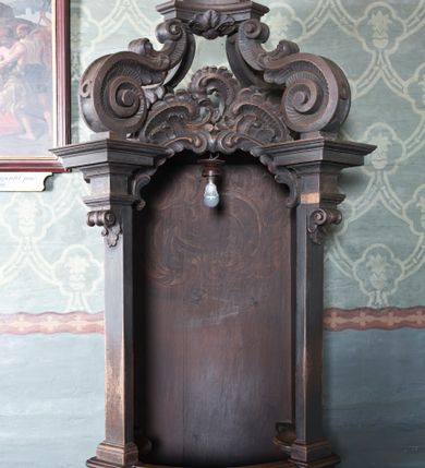 Zdjęcie nr 1: Konfesjonał drewniany o formach architektonicznych, na rzucie zbliżonym do prostokąta o wklęsło-wypukłych bokach. Usytuowany na niewysokim, profilowanym cokole; ujęty ustawionymi ukośnie pilastrami o gładkich trzonach zakończonych akantowymi wolutami dźwigającymi odcinki pełnego belkowania zamkniętego wydatnym, profilowanym gzymsem. W zwieńczeniu ażurowa struktura utworzona z  płaskorzeźbionych rocaillów, a powyżej wazon z girlandą ustawiony na postumencie wspartym na czterech wydatnych wolutach dekorowanych dodatkowo cekinami i akantem. Przedpiersie jednodrzwiczkowe z dekoracją płycinową; na zaplecku malowany rocaille. W bocznych ściankach niewielkie płyciny mieszczące ażurowe kratki. Po bokach konfesjonału dostawione klęczniki na rzucie zbliżonym do prostokąta o linii wklęsło-wypukłej.