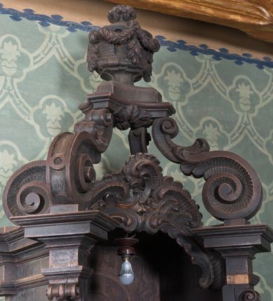 Zdjęcie nr 3: Konfesjonał drewniany o formach architektonicznych, na rzucie zbliżonym do prostokąta o wklęsło-wypukłych bokach. Usytuowany na niewysokim, profilowanym cokole; ujęty ustawionymi ukośnie pilastrami o gładkich trzonach zakończonych akantowymi wolutami dźwigającymi odcinki pełnego belkowania zamkniętego wydatnym, profilowanym gzymsem. W zwieńczeniu ażurowa struktura utworzona z  płaskorzeźbionych rocaillów, a powyżej wazon z girlandą ustawiony na postumencie wspartym na czterech wydatnych wolutach dekorowanych dodatkowo cekinami i akantem. Przedpiersie jednodrzwiczkowe z dekoracją płycinową; na zaplecku malowany rocaille. W bocznych ściankach niewielkie płyciny mieszczące ażurowe kratki. Po bokach konfesjonału dostawione klęczniki na rzucie zbliżonym do prostokąta o linii wklęsło-wypukłej.